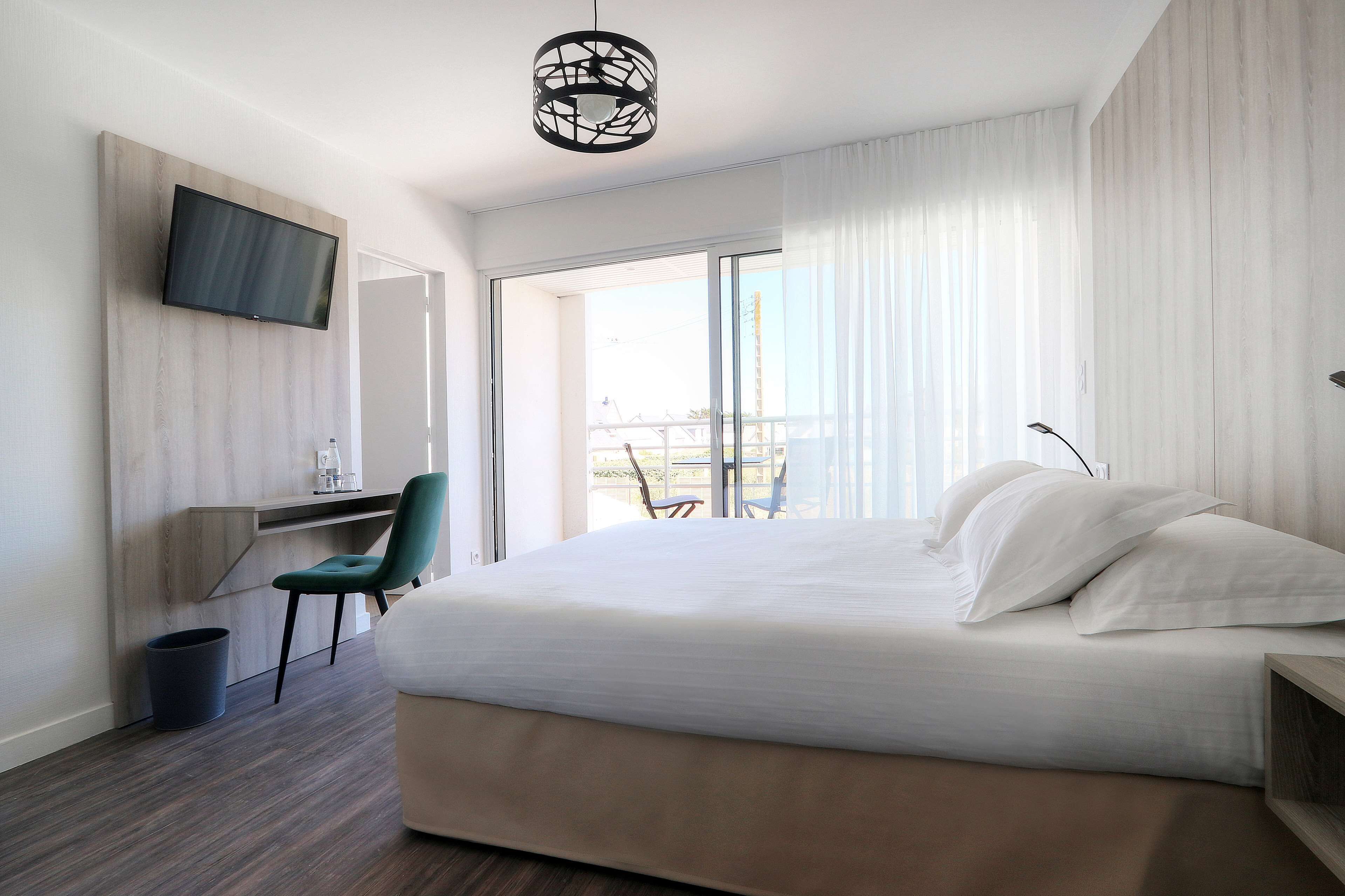 A modern hotel room with a large bed, a desk, a chair, a TV mounted on the wall, and a balcony with a view.