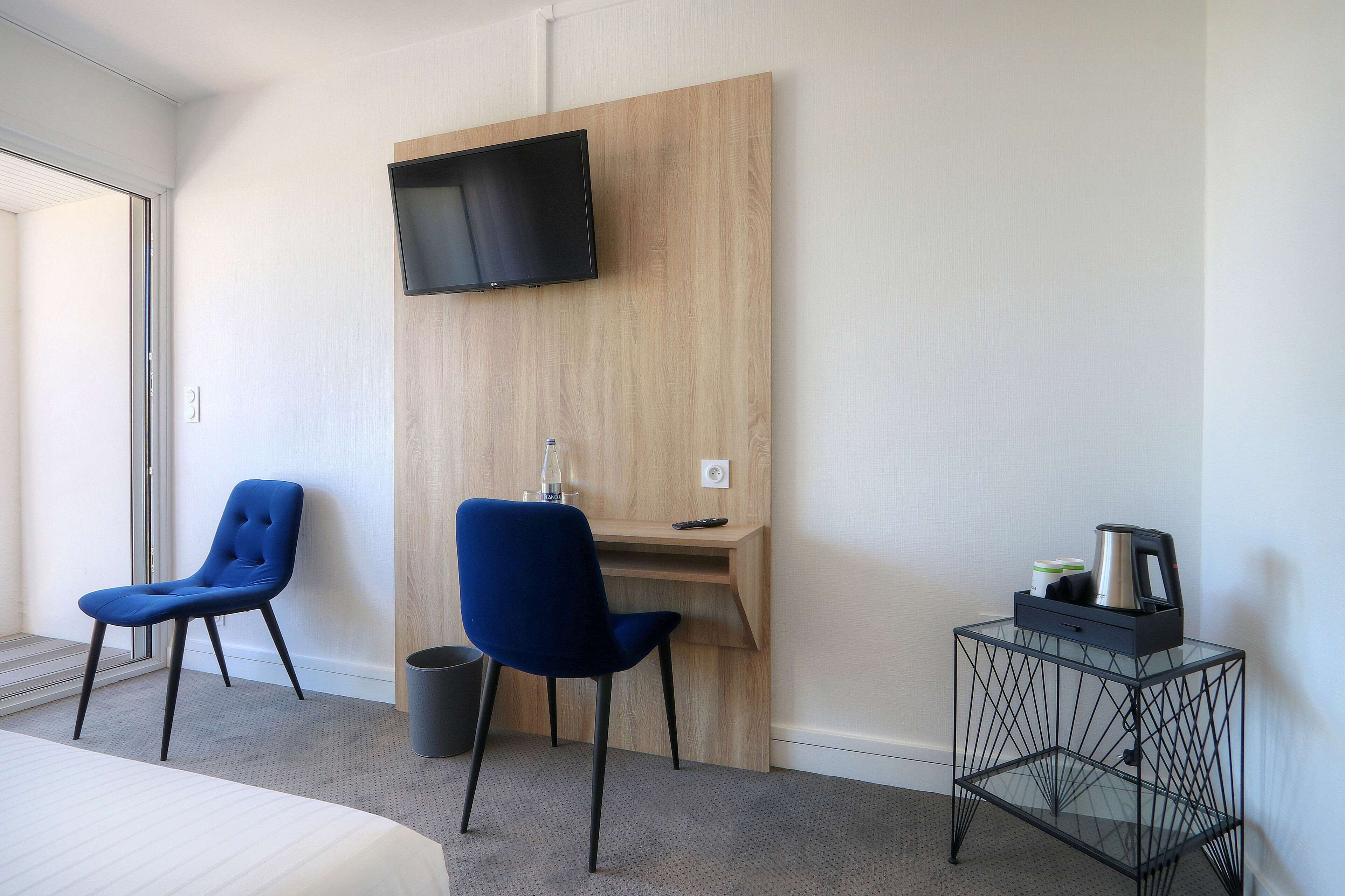 Habitación de hotel con cama, dos sillas azules, un escritorio de madera con televisor, cafetera y cesto de basura.