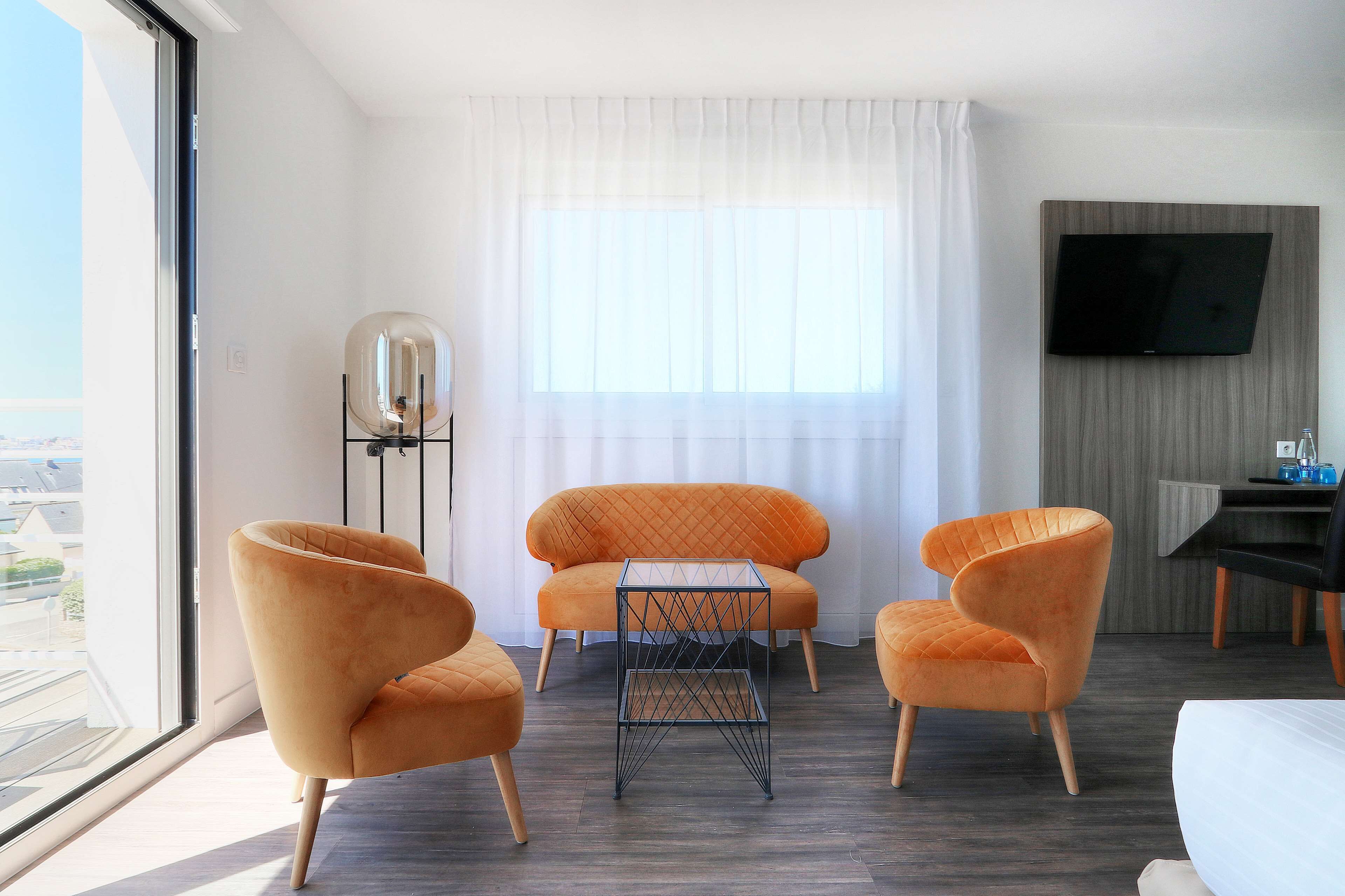 Een moderne hotelkamer met een groot raam, twee oranje fauteuils, een klein bijzettafeltje, een flatscreen televisie aan de muur en uitzicht op de stad.