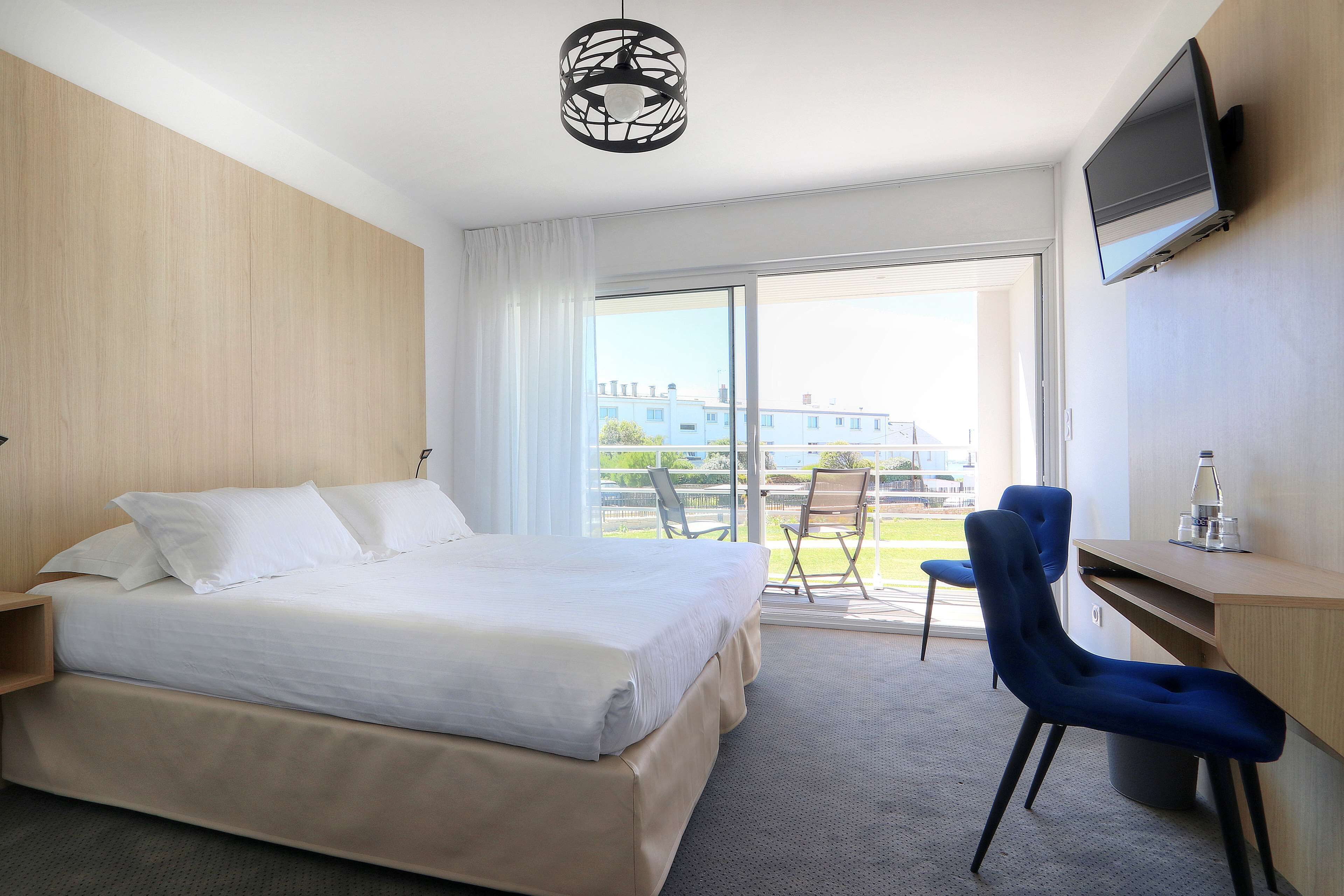 A hotel room with a bed, a desk, a chair, a TV, and a balcony with a view of buildings.