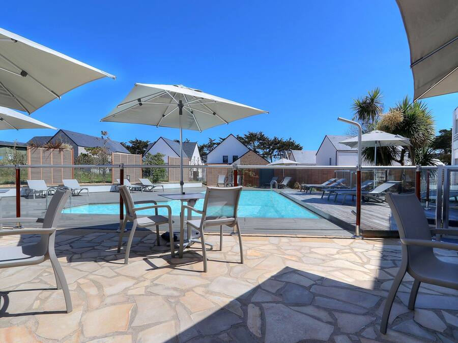 A poolside area with lounge chairs and umbrellas, a stone patio, and a clear view of the pool and surrounding buildings.
