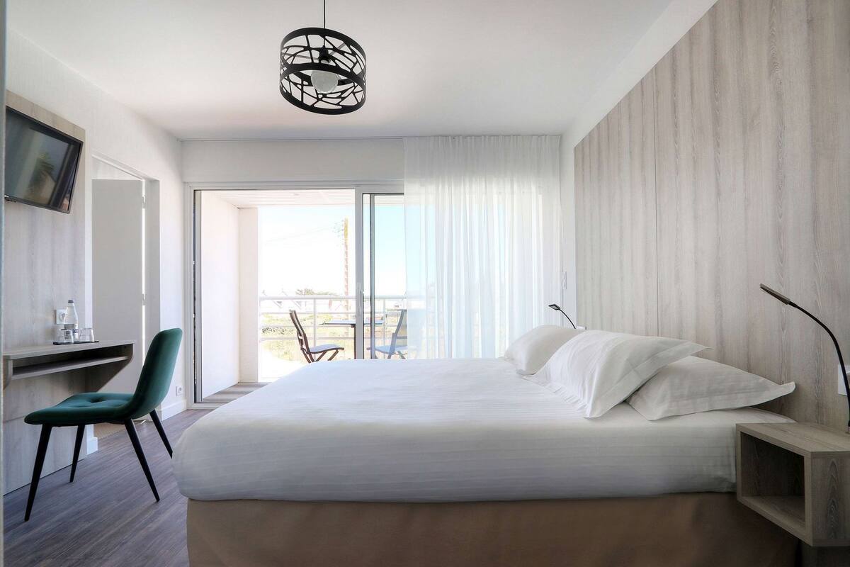 Quarto de hotel moderno com cama, escrivaninha, cadeira e varanda com vista.