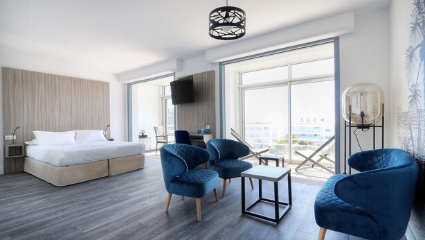 A modern hotel room with a bed, two blue armchairs, a small table, a TV, and large windows offering a city view. - Best Western Hotel Le Bellevue (Quiberon)