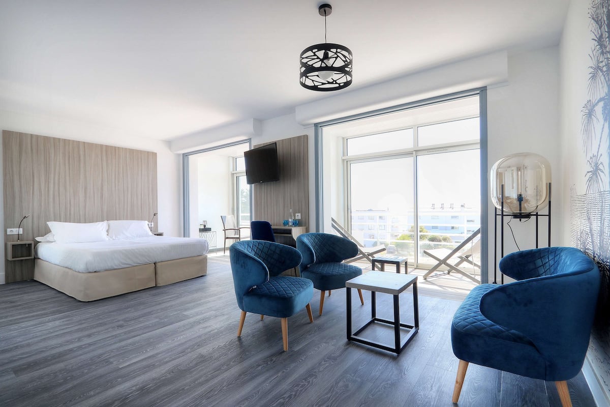 Quarto de hotel moderno com uma cama, duas poltronas azuis, uma pequena mesa, televisão e janelas amplas com vista para a cidade.
