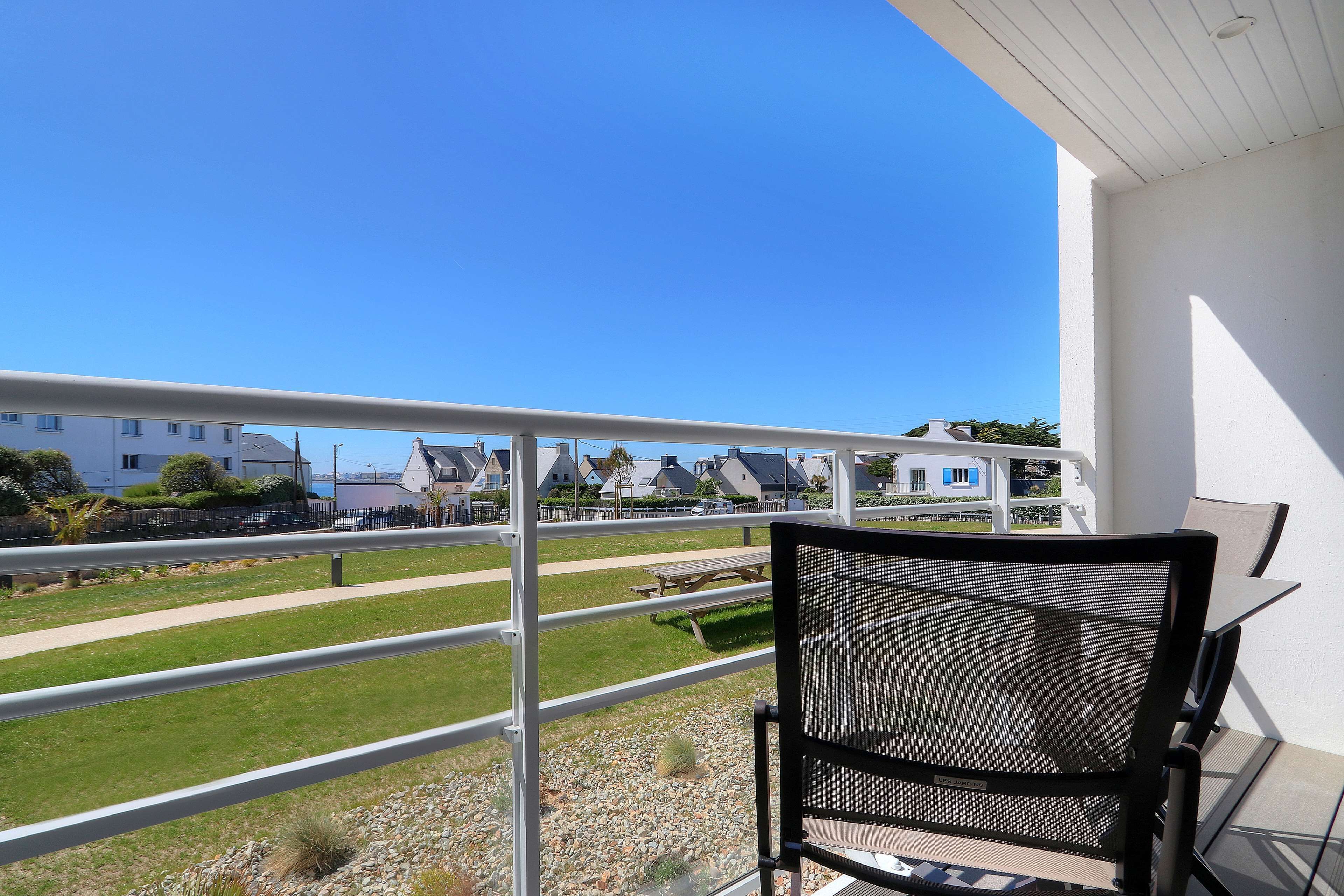 Un balcon doté d’une balustrade en métal, d’une chaise en treillis noir et offrant une vue sur une pelouse bien entretenue et des bâtiments en arrière-plan.