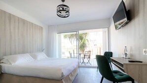 A hotel room with a bed, a desk, a chair, a TV, and a balcony with a view of palm trees. - Best Western Hotel Le Bellevue (Quiberon)