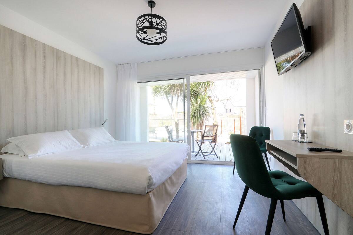 Quarto de hotel com cama, escrivaninha, cadeira, televisão e varanda com vista para palmeiras.