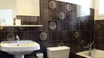 Une salle de bain équipée d’un lavabo, de toilettes et d’une baignoire, avec des carreaux à motifs foncés et un porte-serviettes blanc.