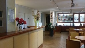 A reception area with a wooden counter, a desk, and chairs. There are potted plants and a bookshelf with books and decorative items. - Hôtel Princess (Vernet-les-Bains)