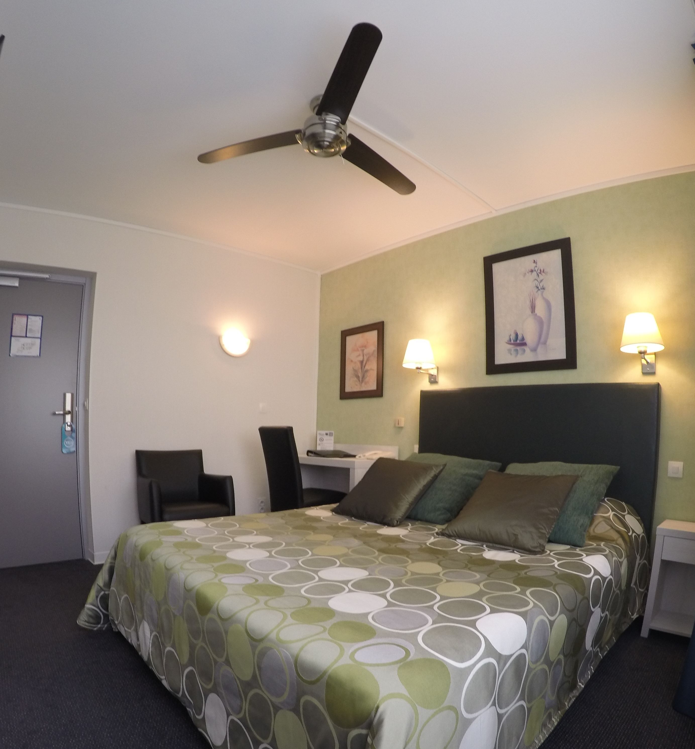 Una habitación de hotel con cama, dos sillones, un escritorio, un ventilador de techo y dos lámparas de pared.
