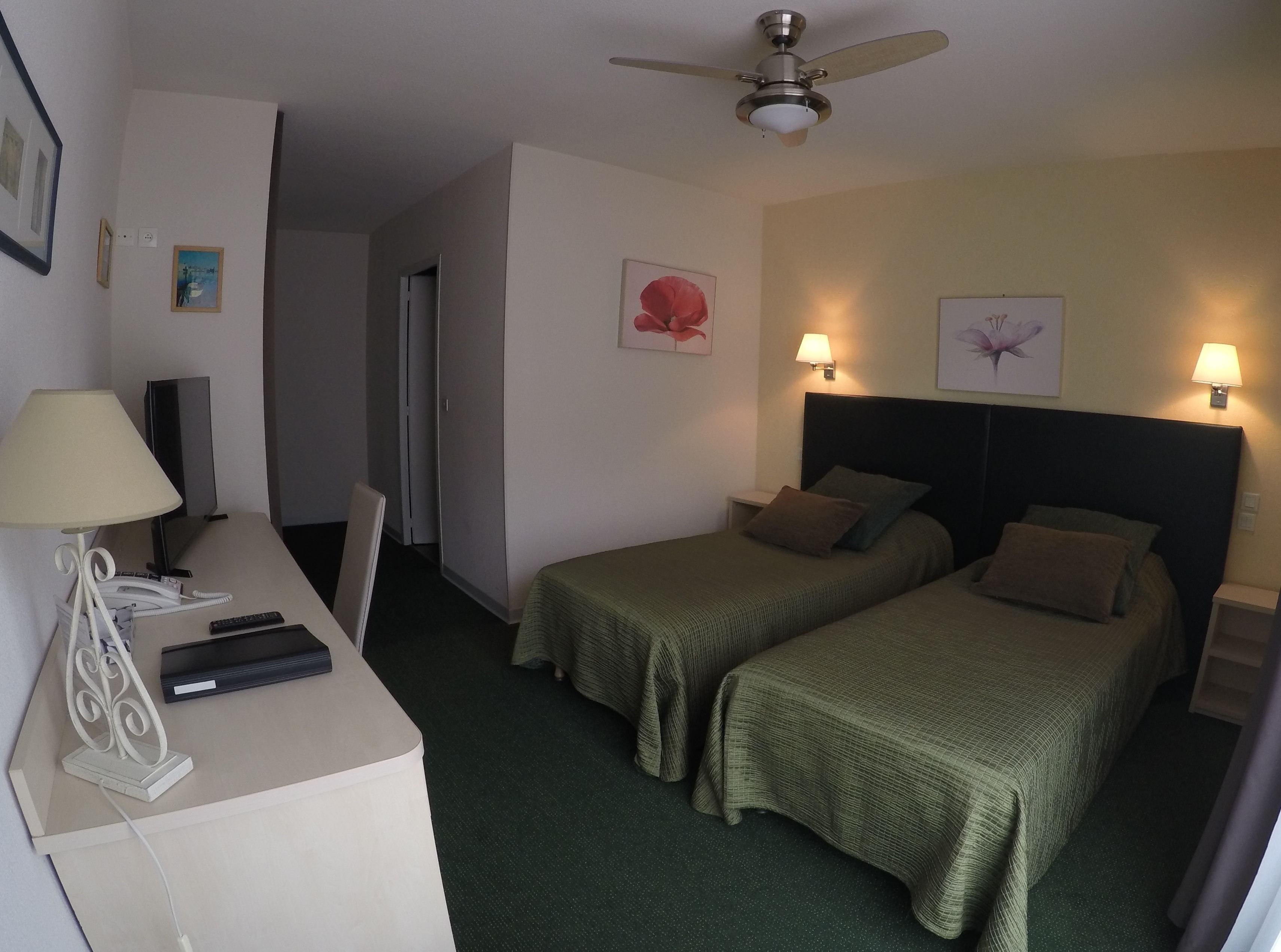 Une chambre d’hôtel avec deux lits simples, un bureau avec une lampe, un téléphone et un ventilateur de plafond.