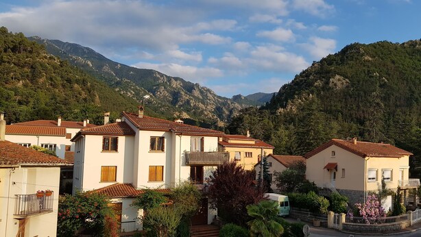 A residential area with houses featuring red-tiled roofs and white walls, surrounded by lush greenery and mountains in the background. - Hôtel Princess (Vernet-les-Bains)