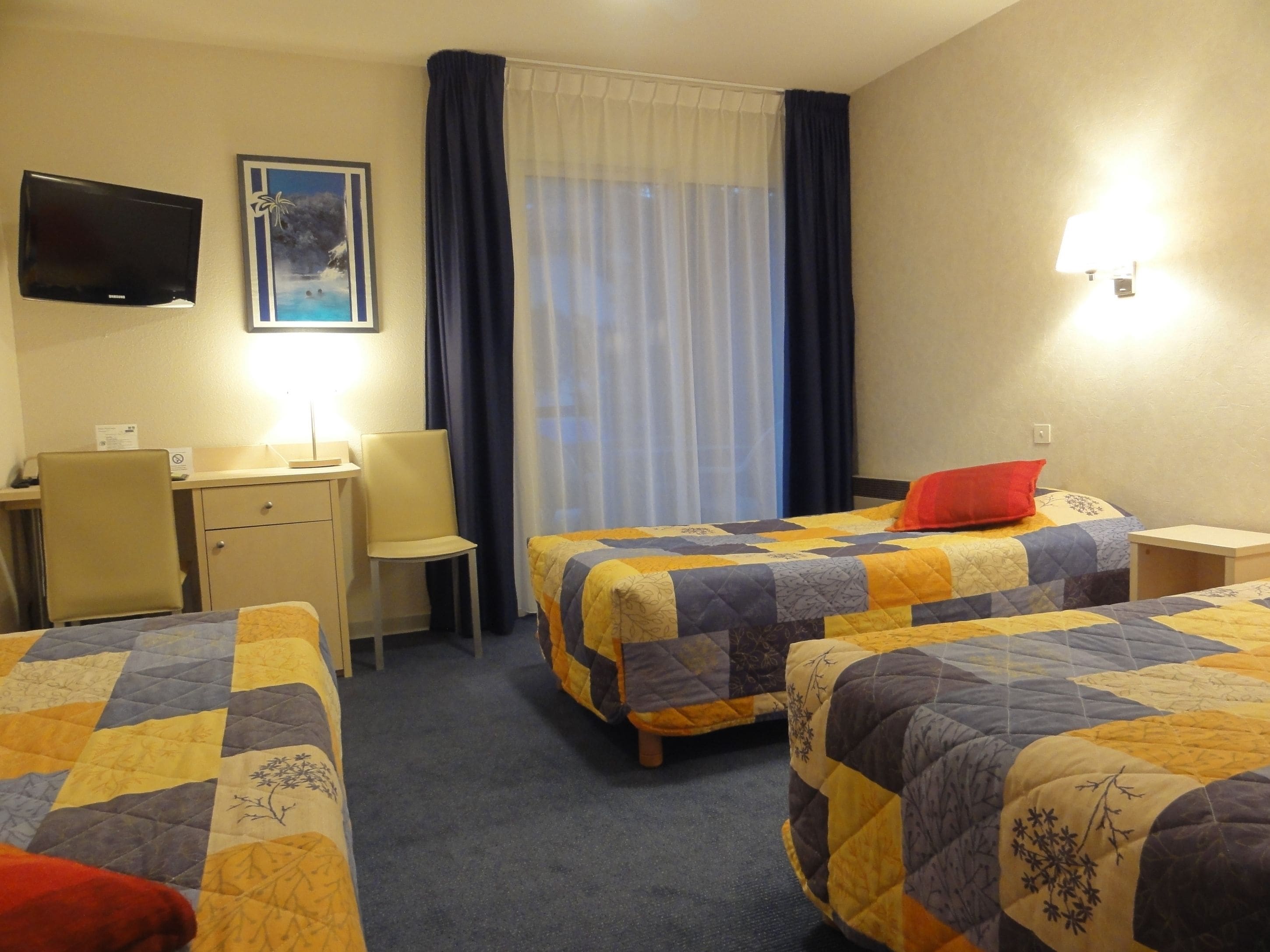 Une chambre d’hôtel avec deux lits, un bureau, une chaise, une télévision et une fenêtre avec des rideaux.
