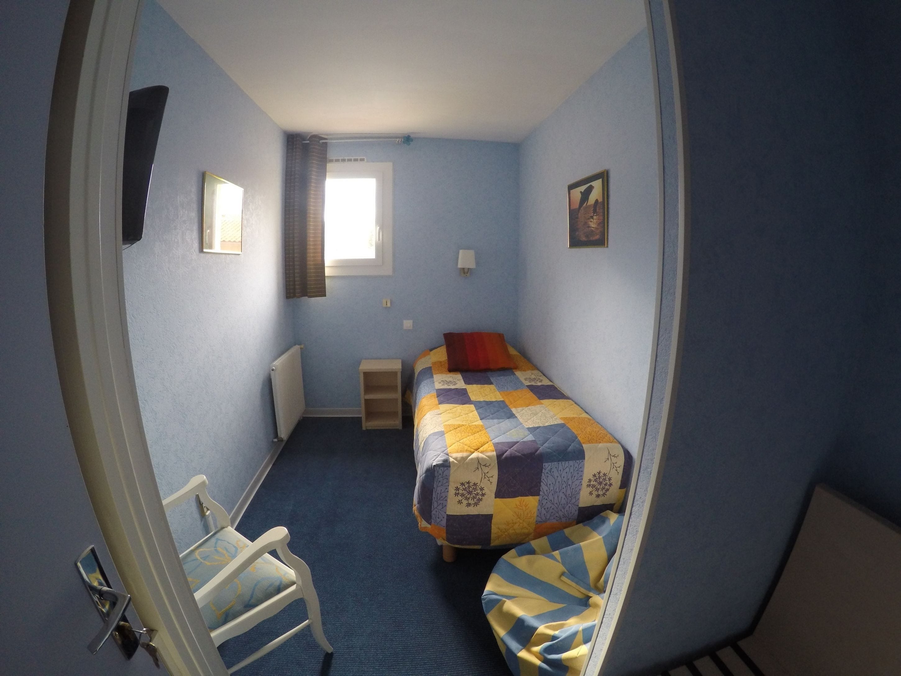 Une petite chambre avec un lit simple, un tapis bleu, une fenêtre avec des rideaux, une table de chevet et une chaise.
