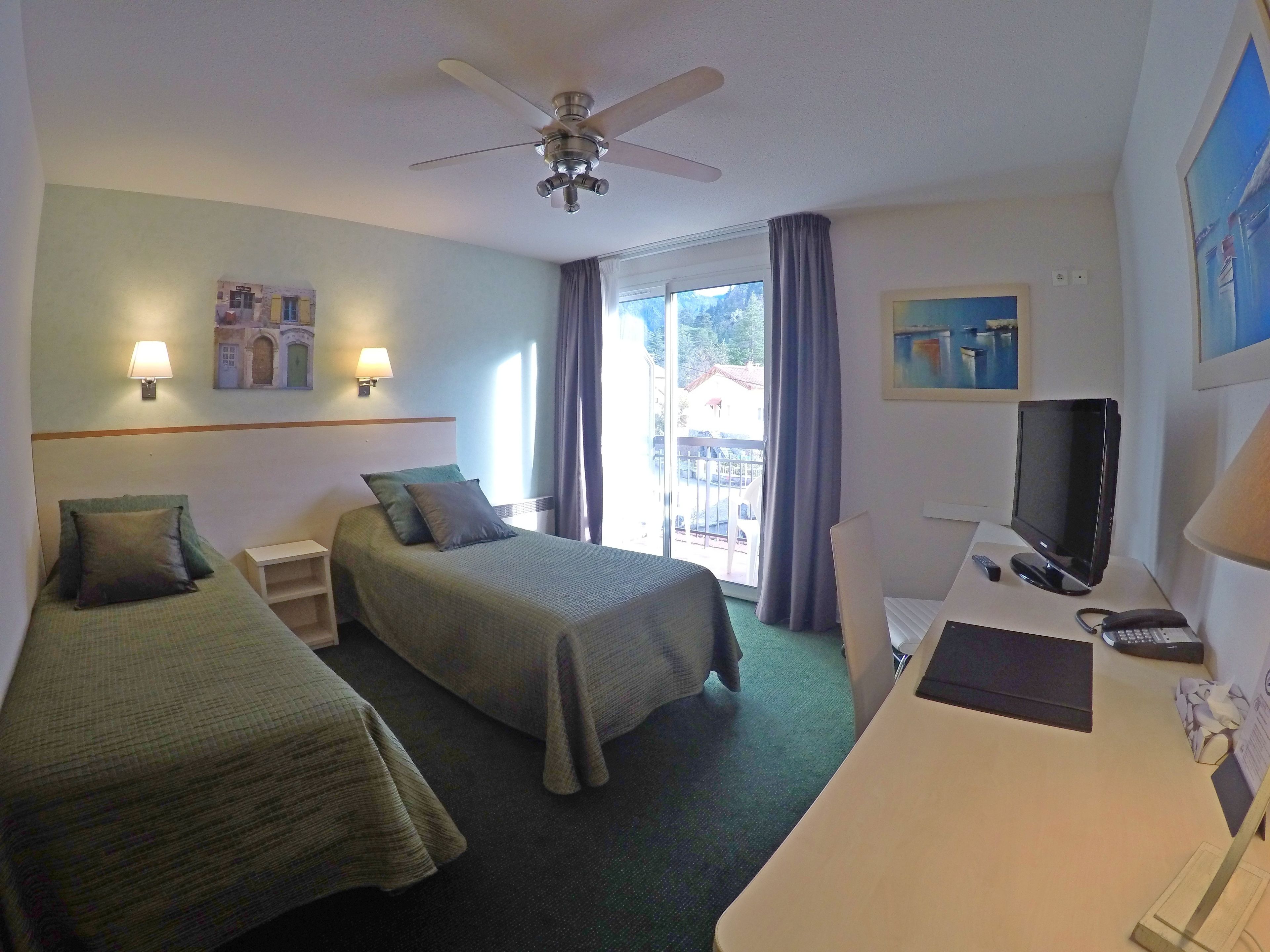 Een hotelkamer met twee bedden, een bureau met een computer, een televisie en een plafondventilator.