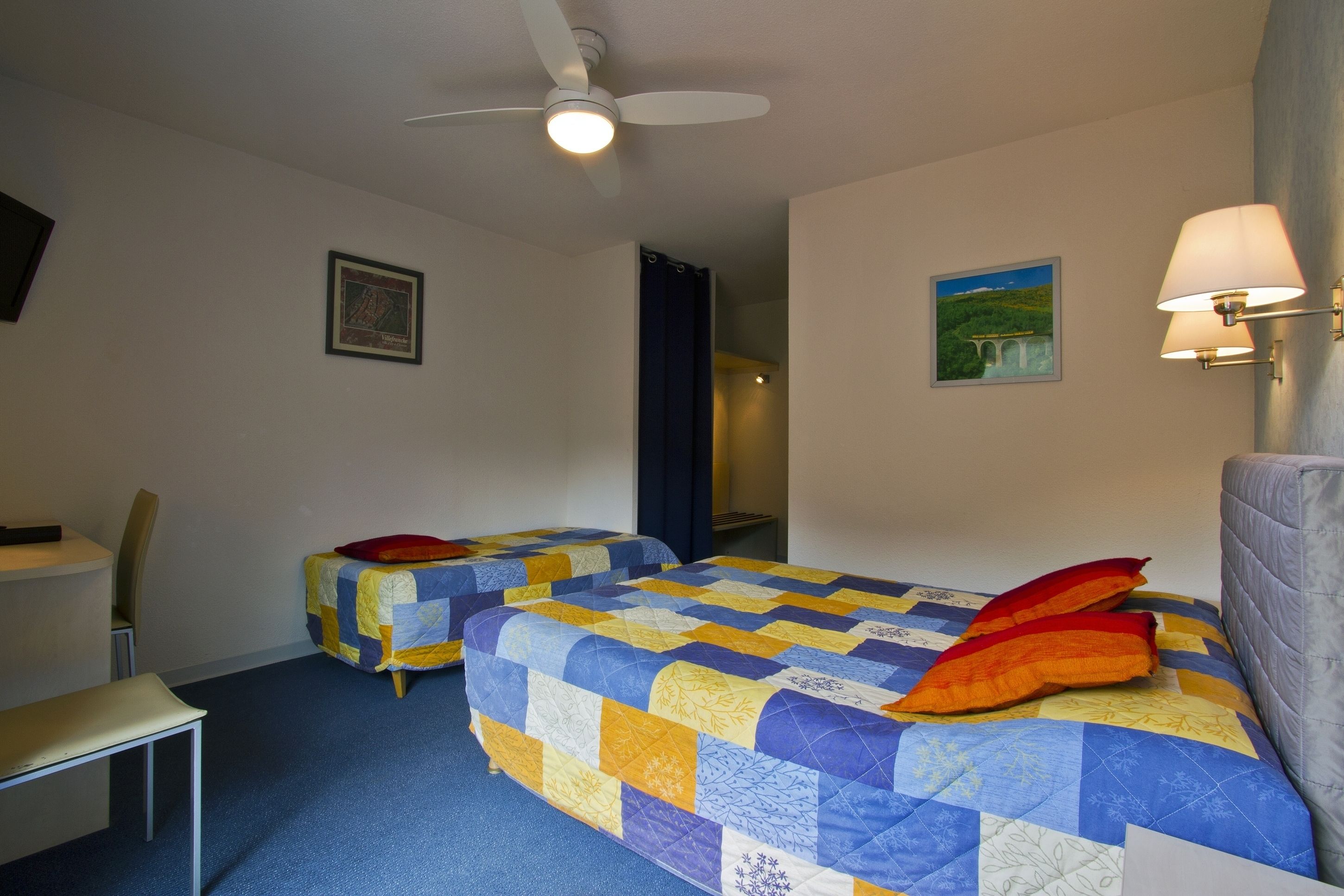 A hotel room with two beds, a desk, a chair, a ceiling fan, and a wall-mounted lamp.