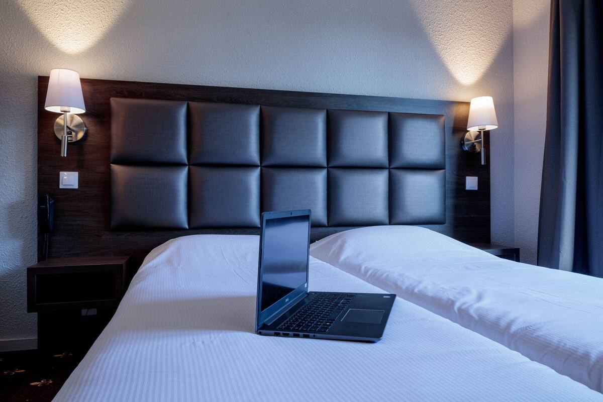 A hotel room with a bed, a laptop on the bed, two bedside lamps, and a dark headboard.