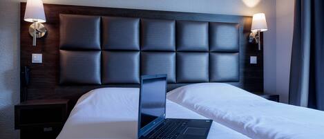 Habitación de hotel con una cama, una laptop sobre la cama, dos lámparas de noche y un cabecero oscuro.
