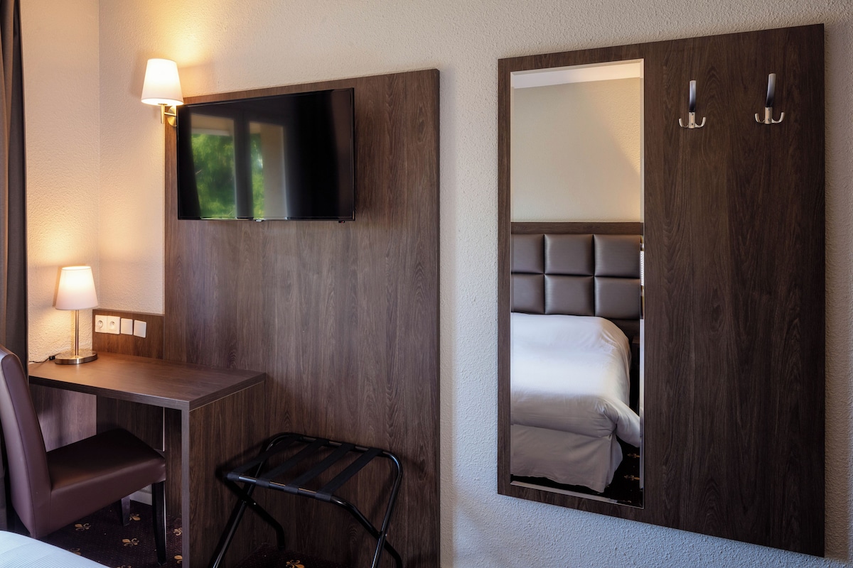 Habitación de hotel con cama, escritorio con silla, televisor montado en la pared y un espejo que refleja la cama.