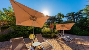 A patio area with sun loungers and large umbrellas, surrounded by greenery. -  Logis Hotel Noemys Morgon (Villie-Morgon)
