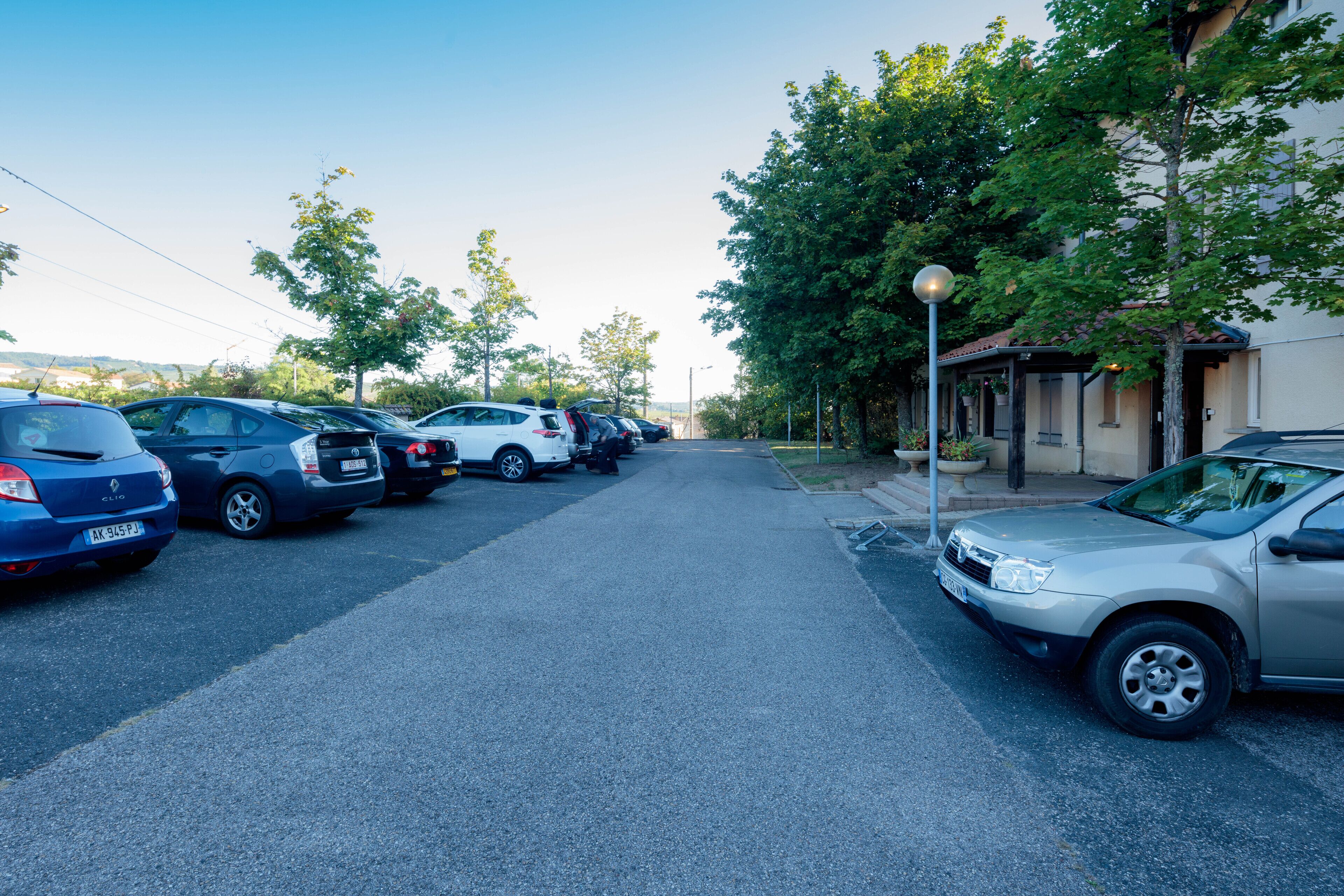 Un parking où plusieurs voitures sont garées le long d’une rue bordée d’arbres, et un bâtiment doté d’une entrée abritée.