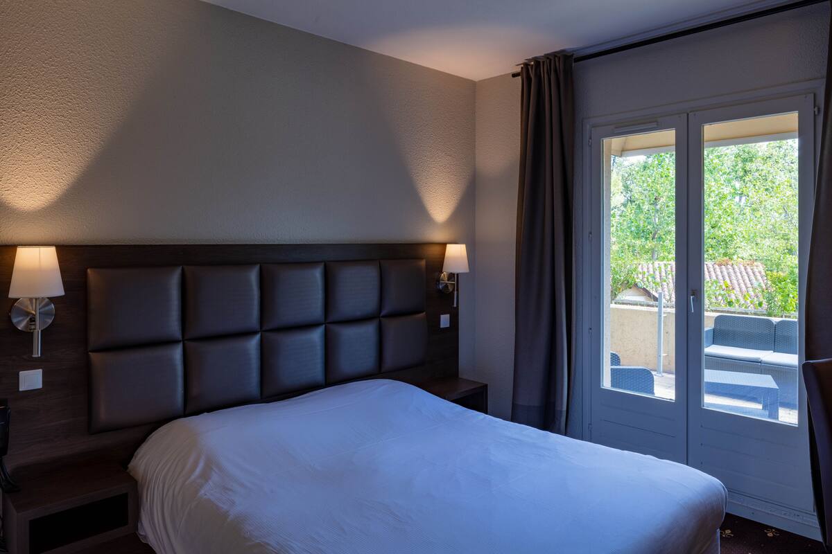 A hotel room with a large bed, two bedside lamps, a headboard, and a view of the outdoors through a sliding door.