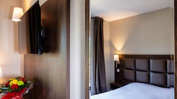 Habitación de hotel con una cama, un televisor montado en la pared, una mesita de noche con lámpara y un ramo de flores sobre una mesita auxiliar.