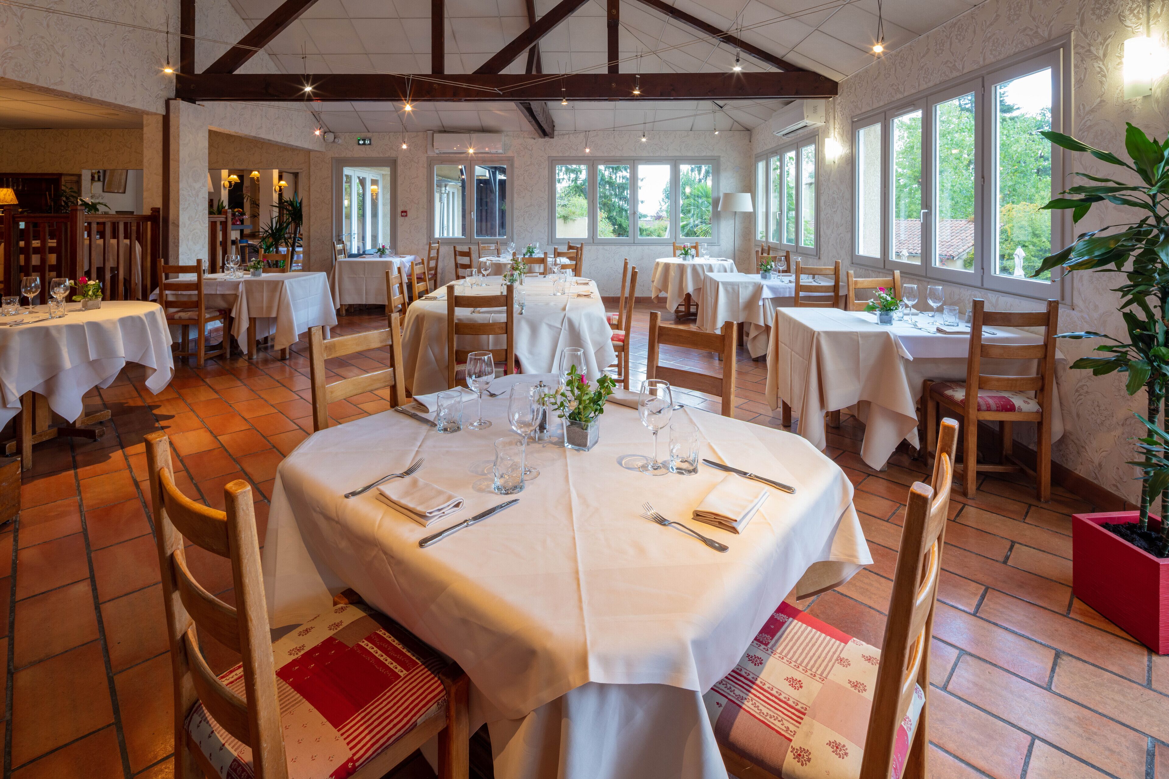 A restaurant with multiple tables set for dining, large windows, and wooden beams on the ceiling.