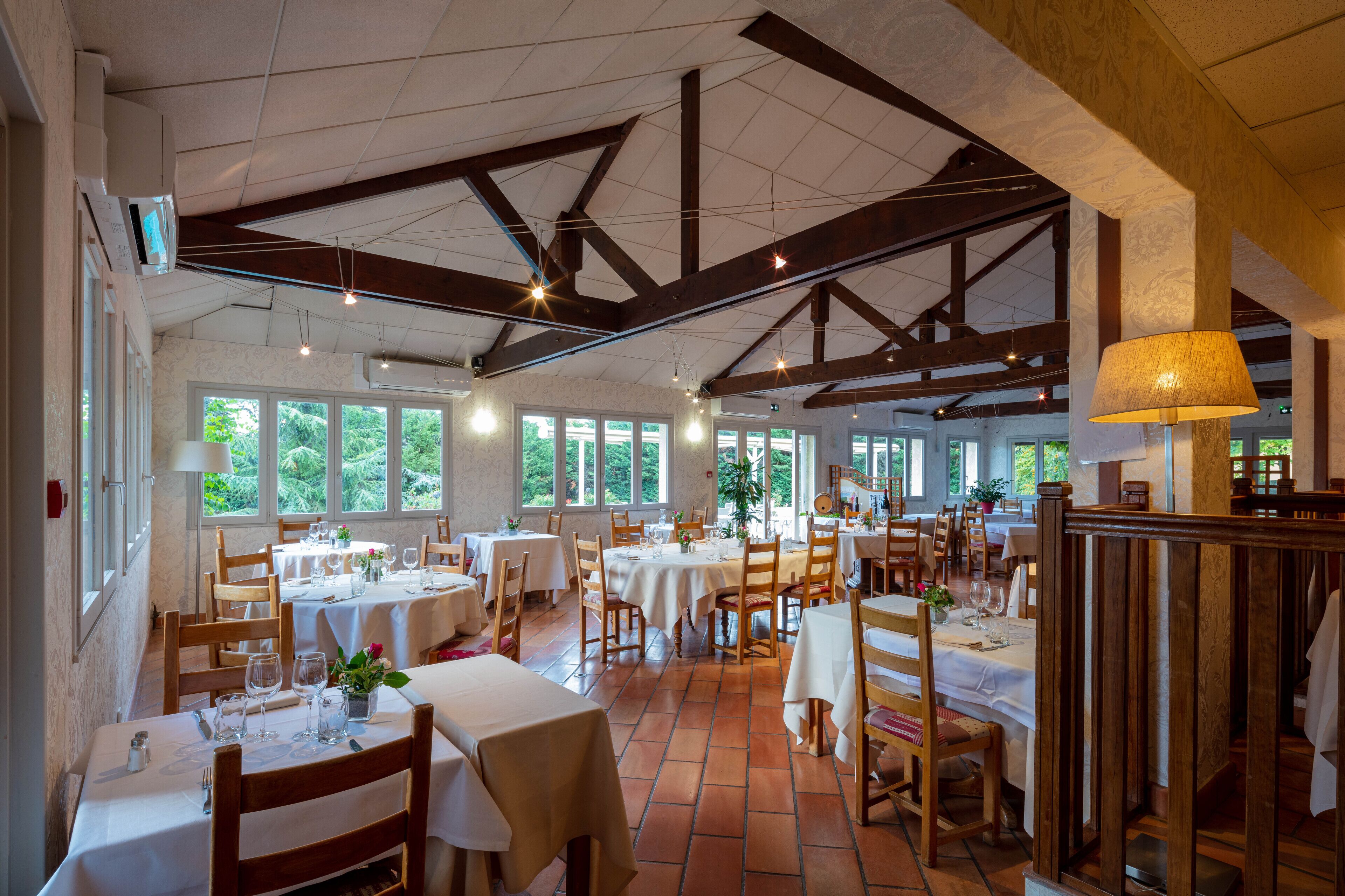 A restaurant with multiple tables and chairs, large windows, and a high ceiling with exposed beams.