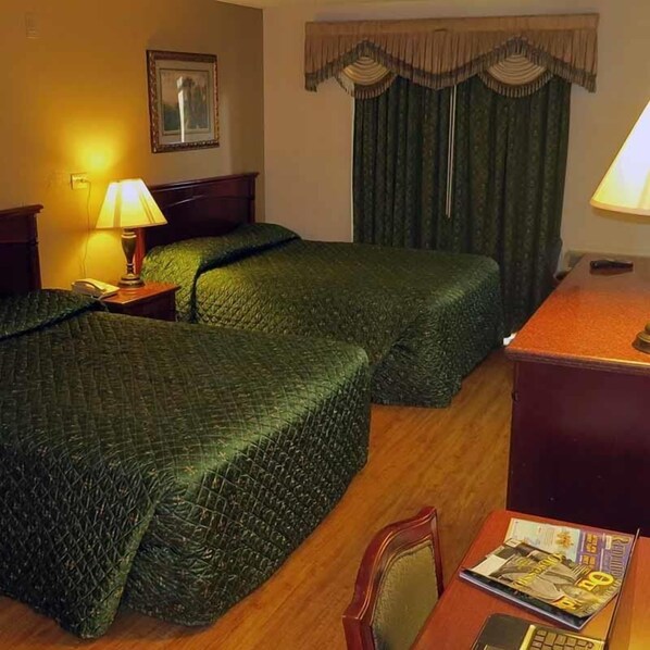 Une chambre d’hôtel avec deux lits, un bureau, une lampe, un tableau encadré au mur et une fenêtre avec des rideaux.