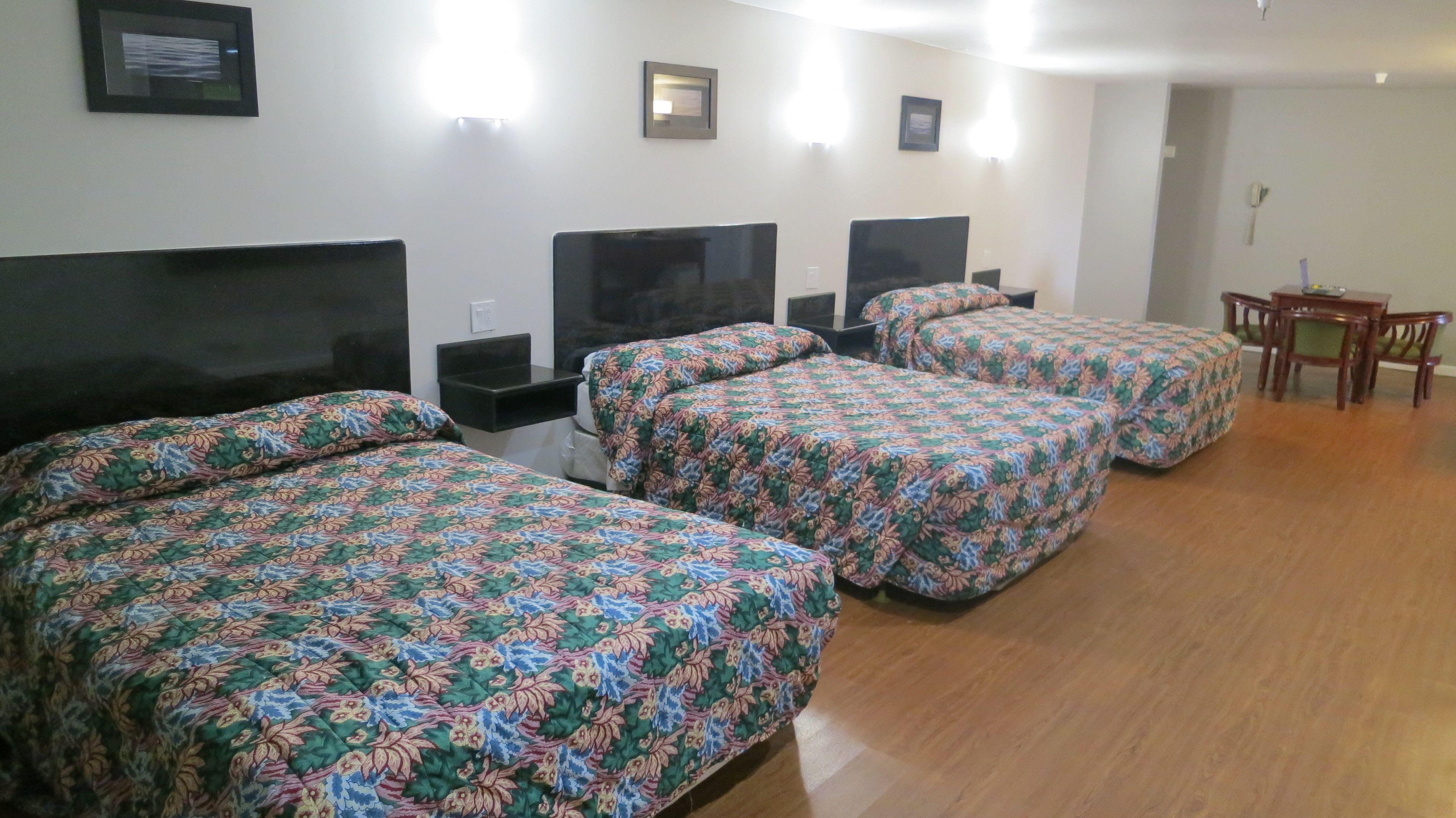 Quarto de hotel com quatro camas, cada uma com colcha estampada em flor. Há uma pequena mesa de madeira e uma cadeira no canto.