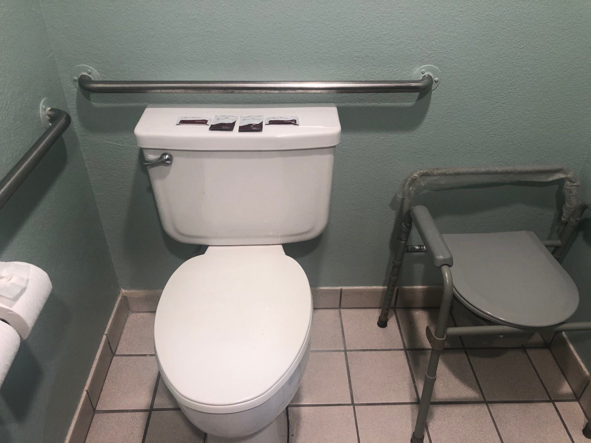 A standard toilet with a raised seat and grab bars, a white toilet paper holder, and a tiled floor.