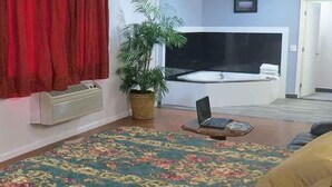 Une pièce avec une grande moquette à motifs floraux, une plante en pot, un ordinateur portable posé sur une petite table et un lit avec un rideau rouge.