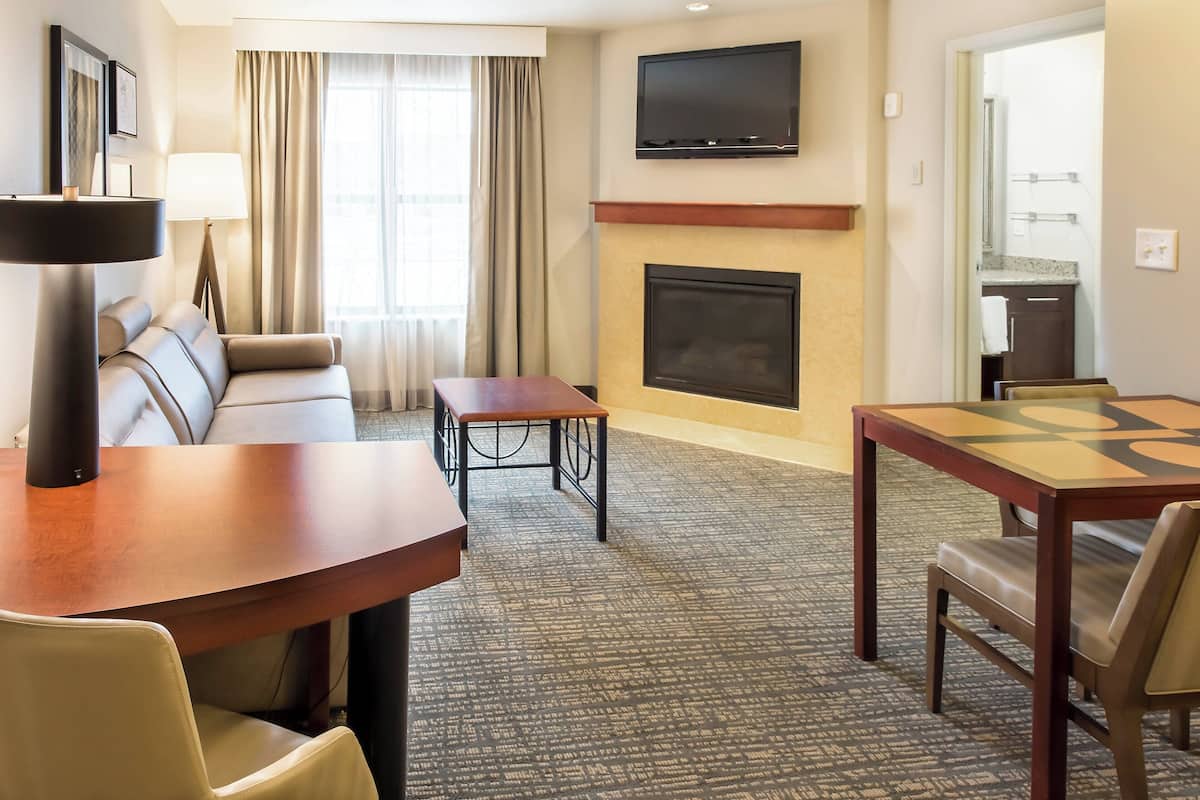 A hotel room with a sofa, a dining table, a TV mounted on the wall, and a fireplace.