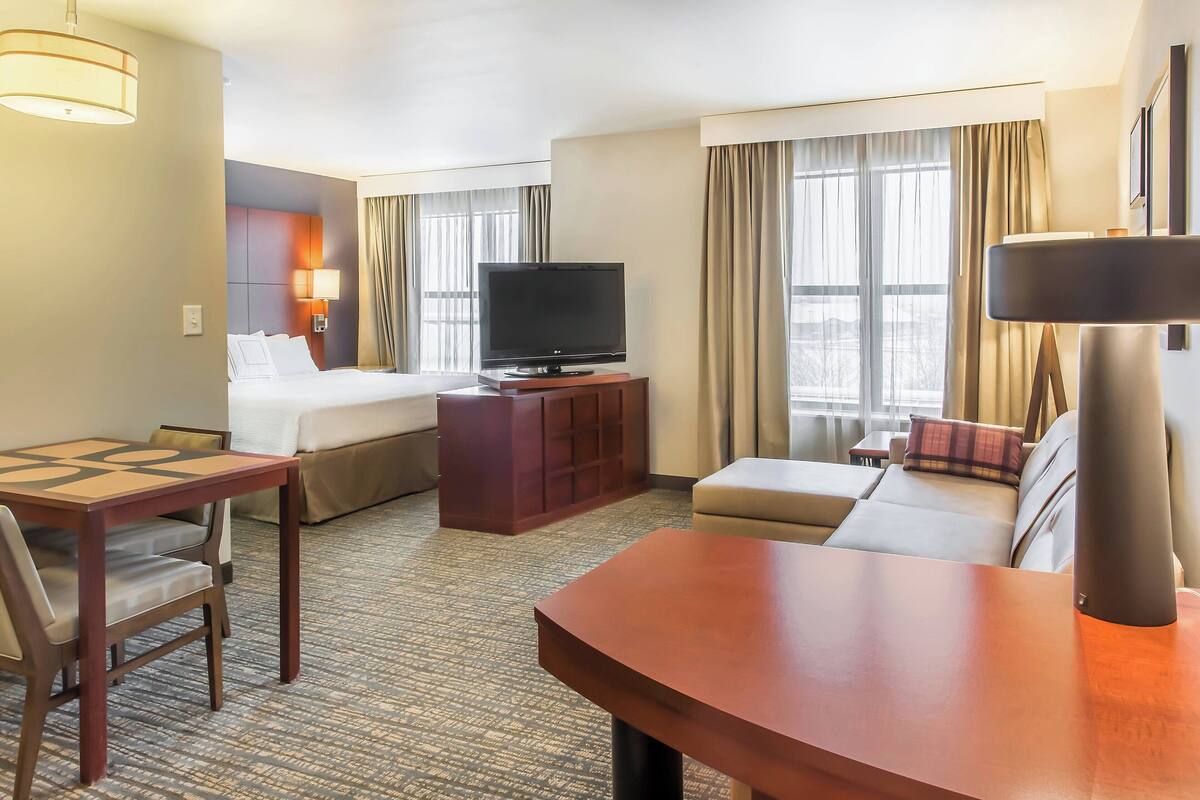 A hotel room with a bed, a TV on a wooden stand, a sofa, a desk with a chair, and a lamp.