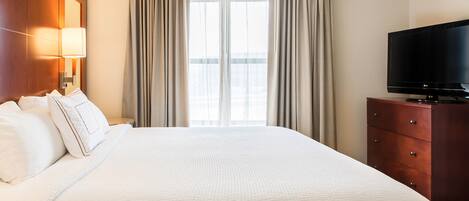 Habitación de hotel con una cama grande, un televisor de pantalla plana, un aparador de madera y un ventanal con cortinas.