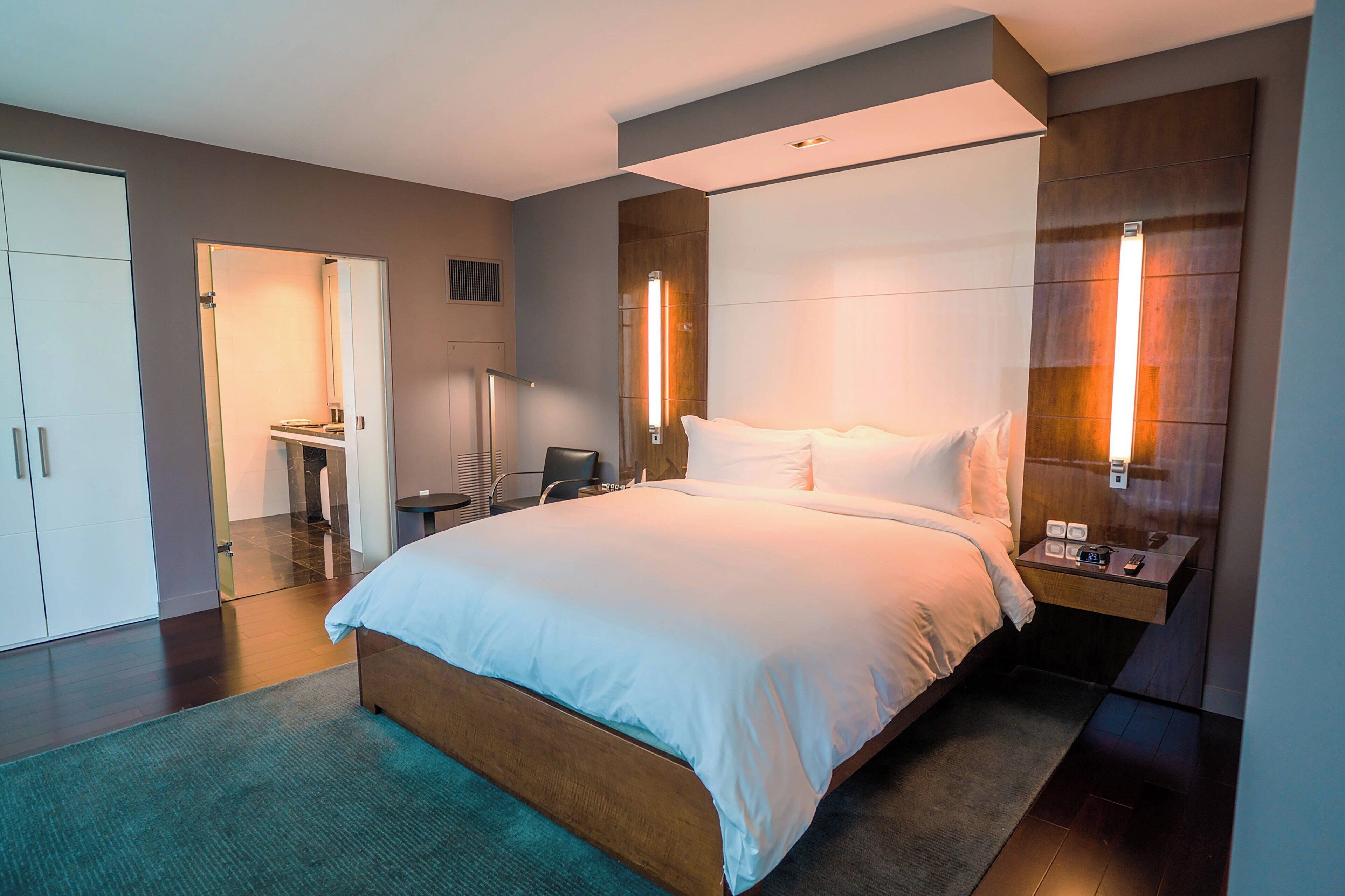A modern hotel room with a large bed, bedside tables, a wardrobe, and a bathroom visible through an open door.
