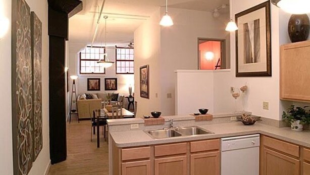 A modern kitchen with wooden cabinets, a white dishwasher, and a dining area with a sofa and table. - Bridgestreet at the Bingham (Cleveland)