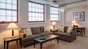 A living room with two couches, a coffee table, and two lamps. There are windows with blinds, a painting on the wall, and a carpeted floor. - Bridgestreet at the Bingham (Cleveland)