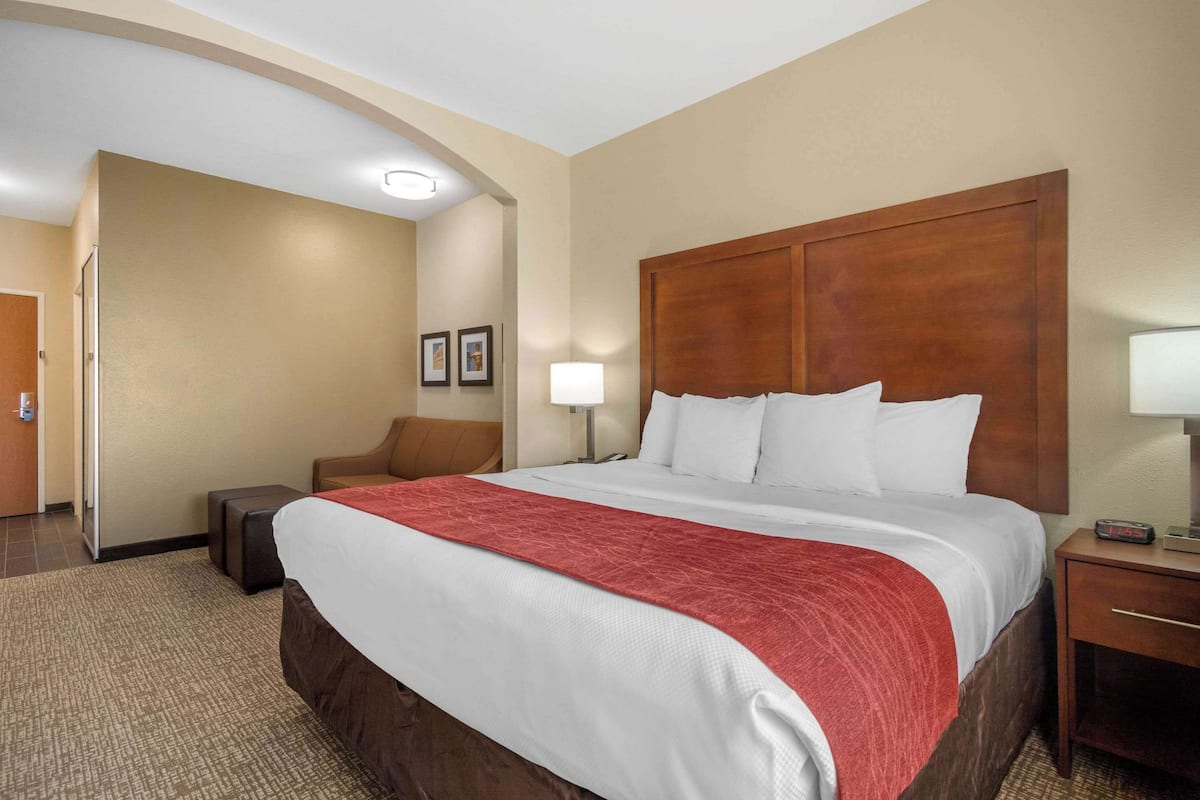 A hotel room with a large bed, a wooden headboard, a bedside table with a lamp, and a small seating area with a chair and ottoman.