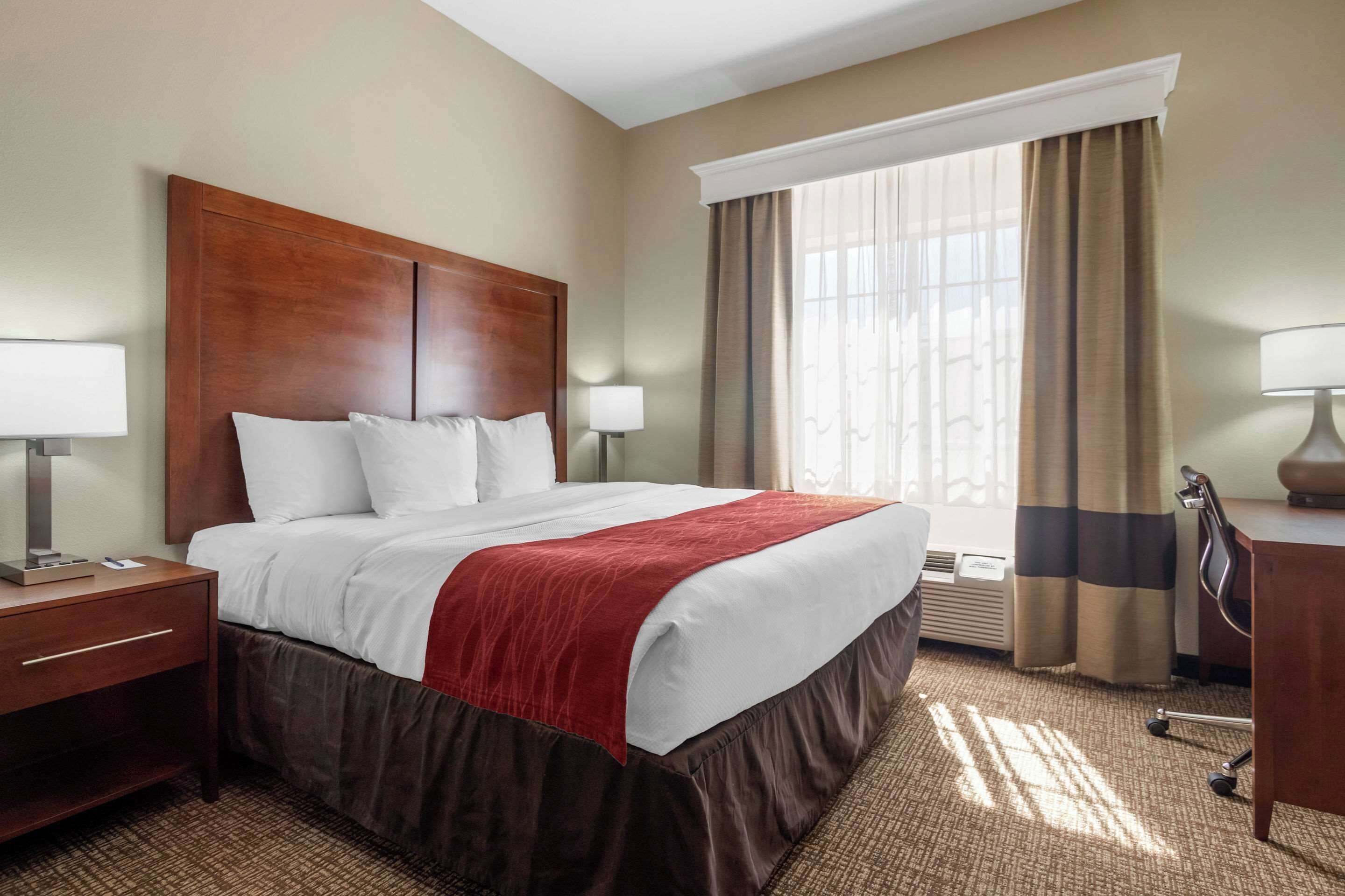 Habitación de hotel con una cama grande, dos mesitas de noche con lámparas, un escritorio con silla y un ventanal con cortinas.