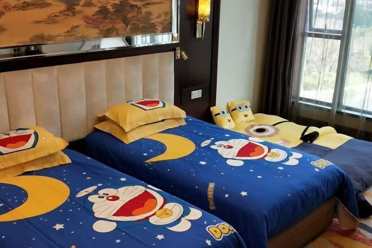 A hotel room with two beds, each with a blue bedspread featuring cartoon characters. There is a window with curtains, a door with a light switch, and a wall-mounted TV.