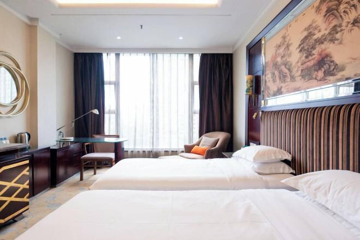 A hotel room with two beds, a desk, a chair, a large window with curtains, and a painting on the wall.