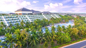 A hotel with multiple balconies, a pool area surrounded by palm trees, and a view of distant mountains. - Sea Links Beach Hotel (Phu Thuy)