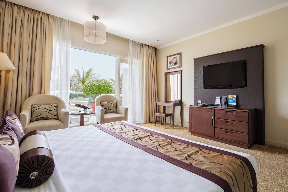 Een hotelkamer met een groot bed, een televisie, een bureau, een stoel en uitzicht op palmbomen.