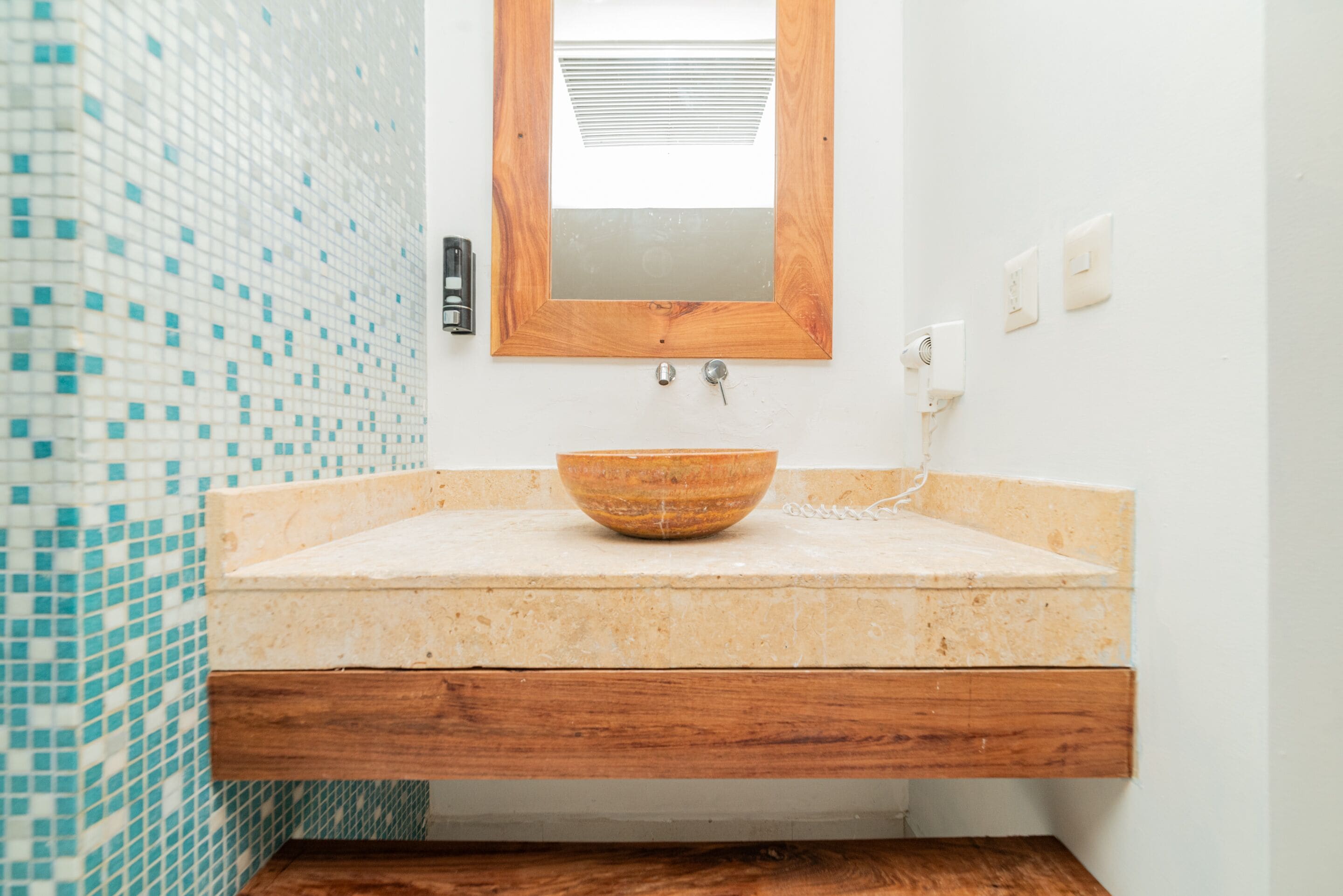 Une salle de bain avec un mur recouvert de mosaïques, un miroir encadré en bois, un lavabo en forme de coupe et une étagère en bois.