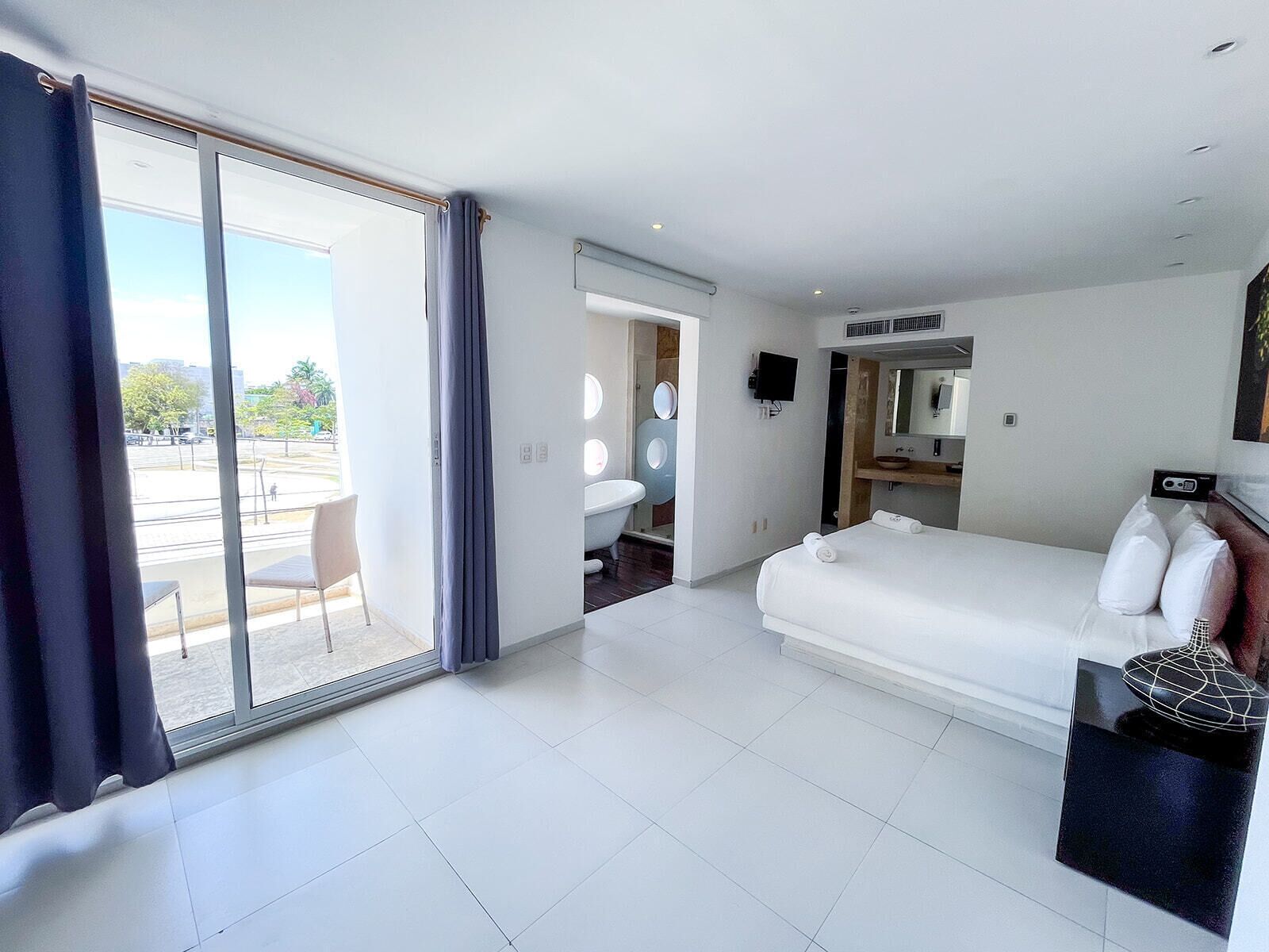 A modern hotel room with a large sliding glass door leading to an outdoor area, a single bed with white linens, a small bathroom with a bathtub, and a flat-screen TV mounted on the wall.