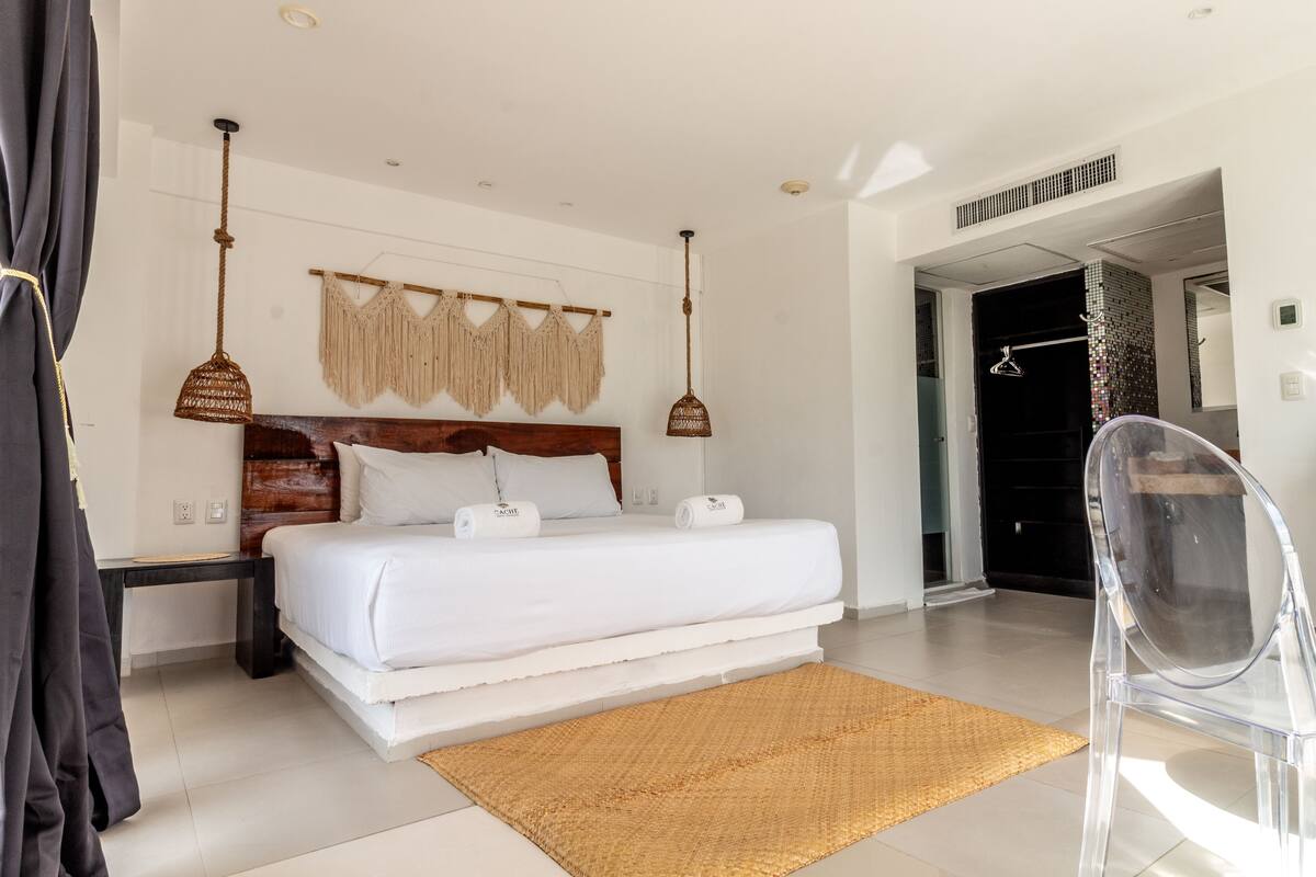 Une chambre d’hôtel moderne avec un grand lit, une tête de lit en bois, un tapis tissé et une chaise transparente.