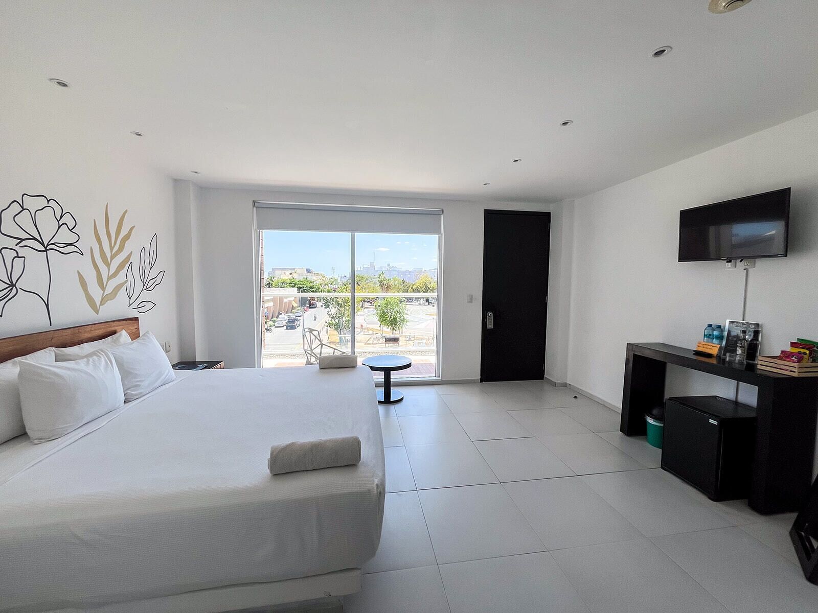 Habitación de hotel moderna con una cama grande, un escritorio con televisor y un balcón con vistas a la ciudad.