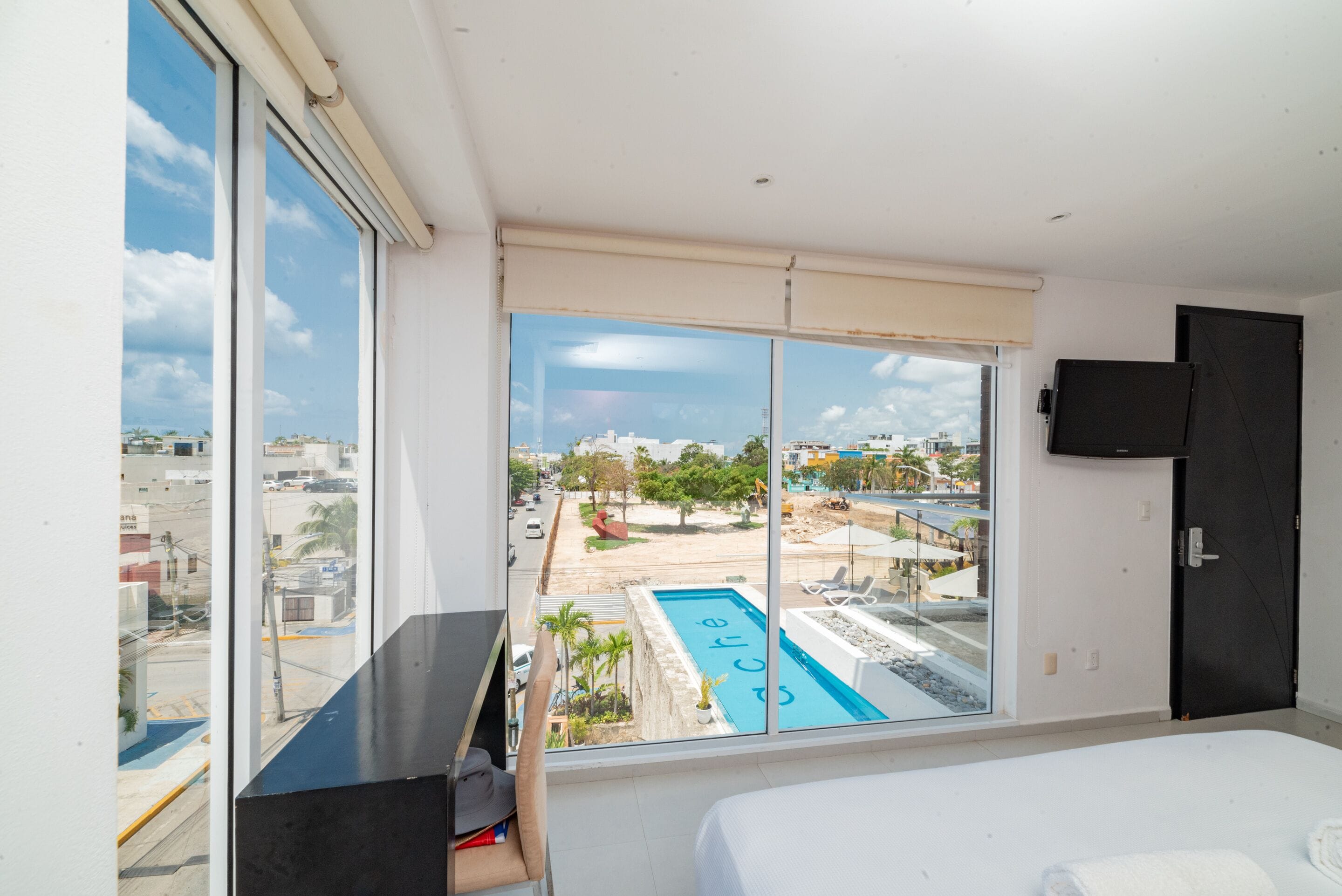 A hotel room with a large window, a bed, a TV, and a view of a swimming pool and beach.